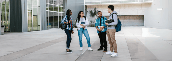 Symbolbild Vier Studierende stehen auf dem Campus beisammen und unterhalten sich.